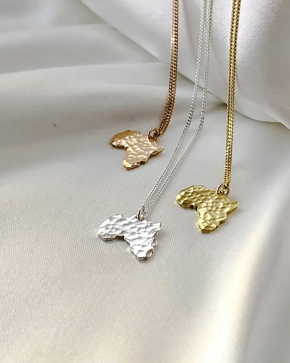Three Africa-shaped pendant necklaces in contrasting metal tones on pale fabric.